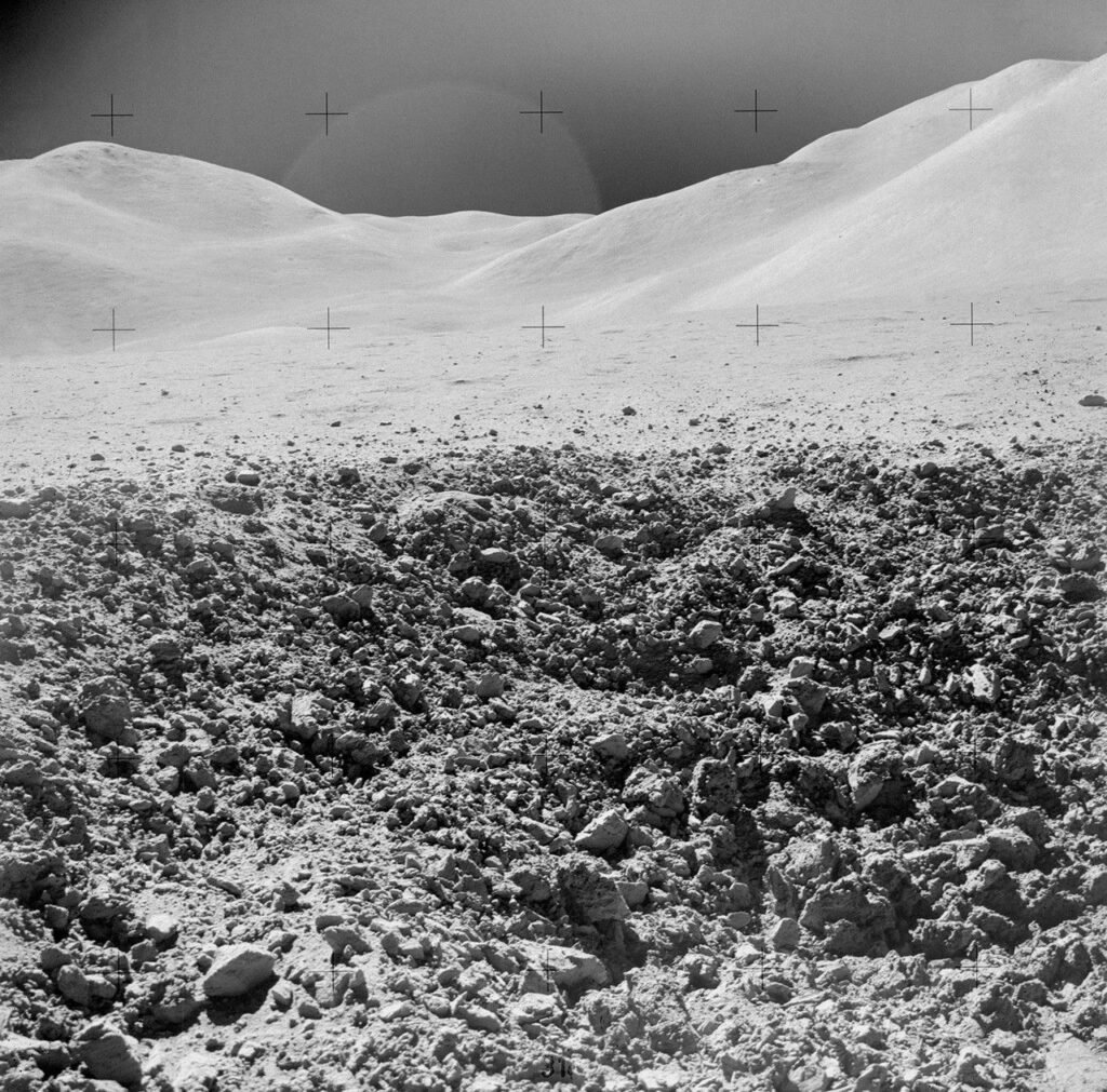 View from surface of lunar crater. The foreground looks like an expanse of rocky rubble. In the background, lighter-colored, dune-shaped hills rise under a dark sky.