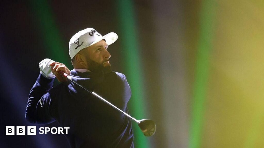 Jon Rahm tees off on the 16th hole on day one of LIV Golf Riyadh. He has a black beard and is wearing a white cap, a white glove on his left hand and a navy sweater.