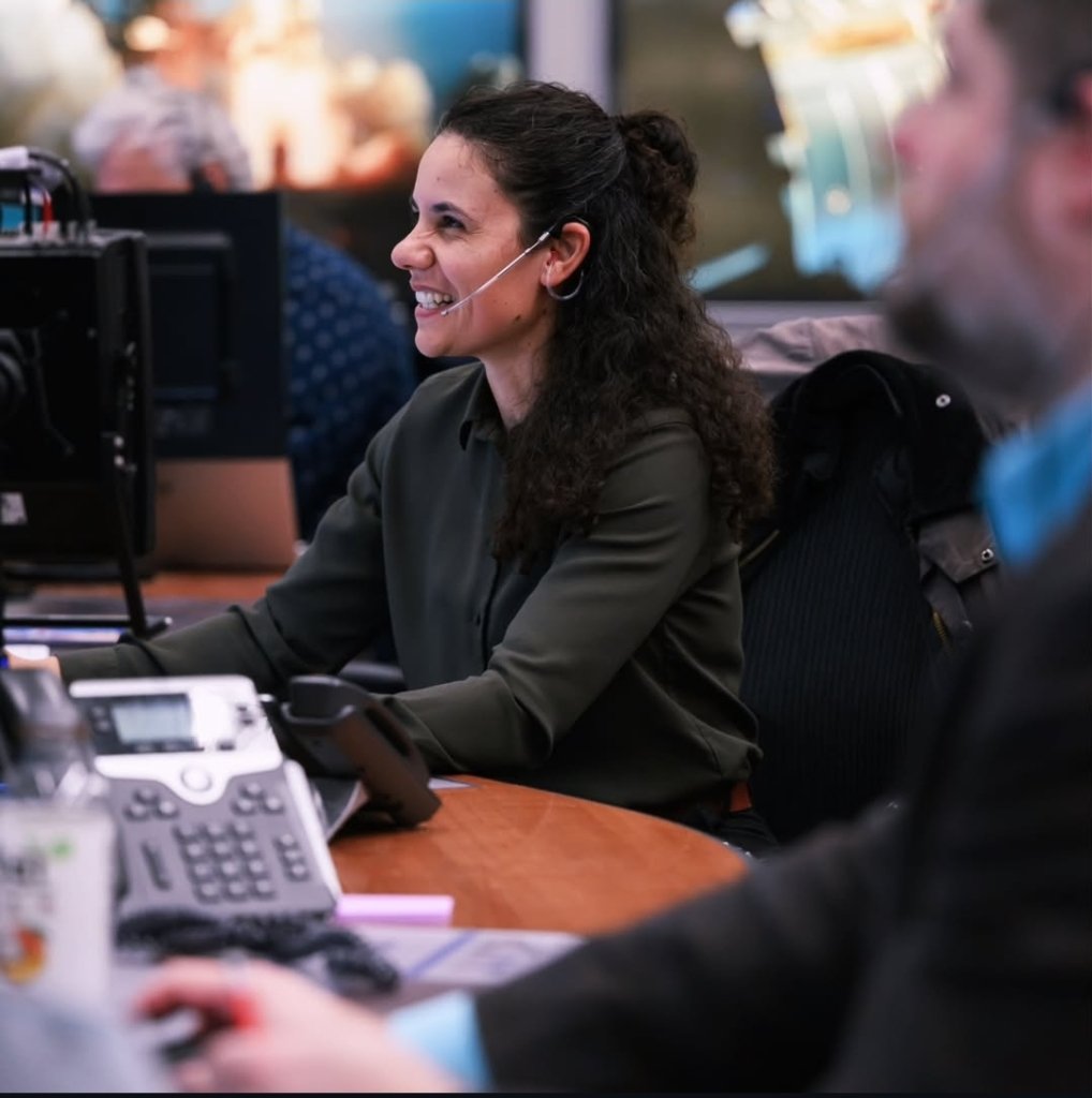 A woman at a desk with a headset on smiles and looks up.