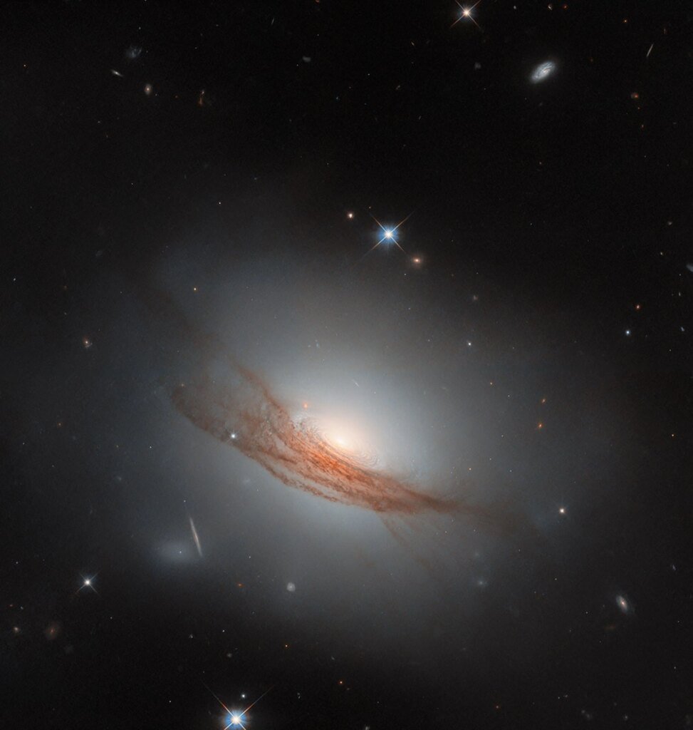 A disk-shaped galaxy. It glows brightly at the center and shines a faint white light all around it. The disk is made up of tightly-packed rings of dust, some darker and some lighter. Wide, long lanes of dark reddish dust cross the galaxy in front of its edge, blocking out some of its light; the long strands twist and break apart at each side. A couple of nearby stars and distant galaxies are also visible on the black background.